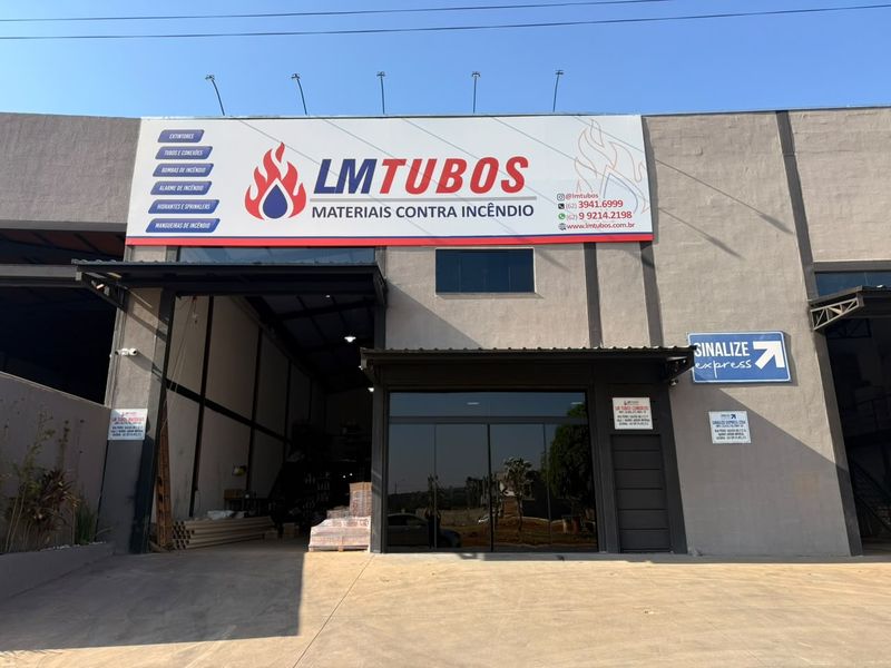 Fachada do galpão da LM Tubos em Goiânia, com o letreiro e o portão aberto mostrando tubos e paletes de material em estoque